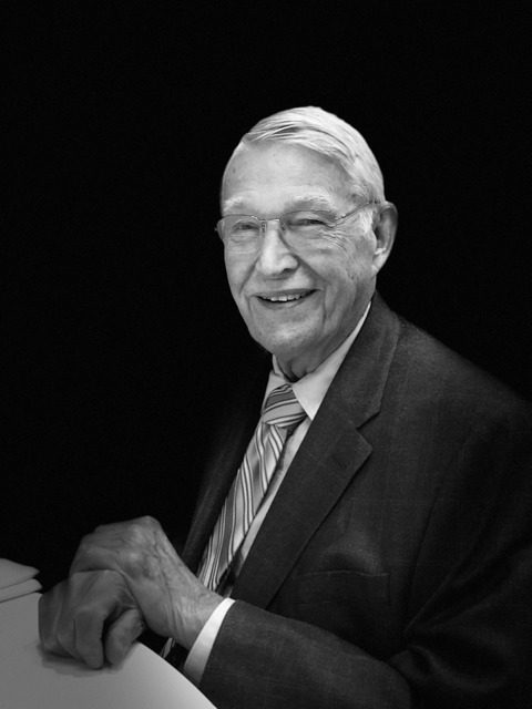 Portrait of an elderly man in a suit smiling warmly against a black background.